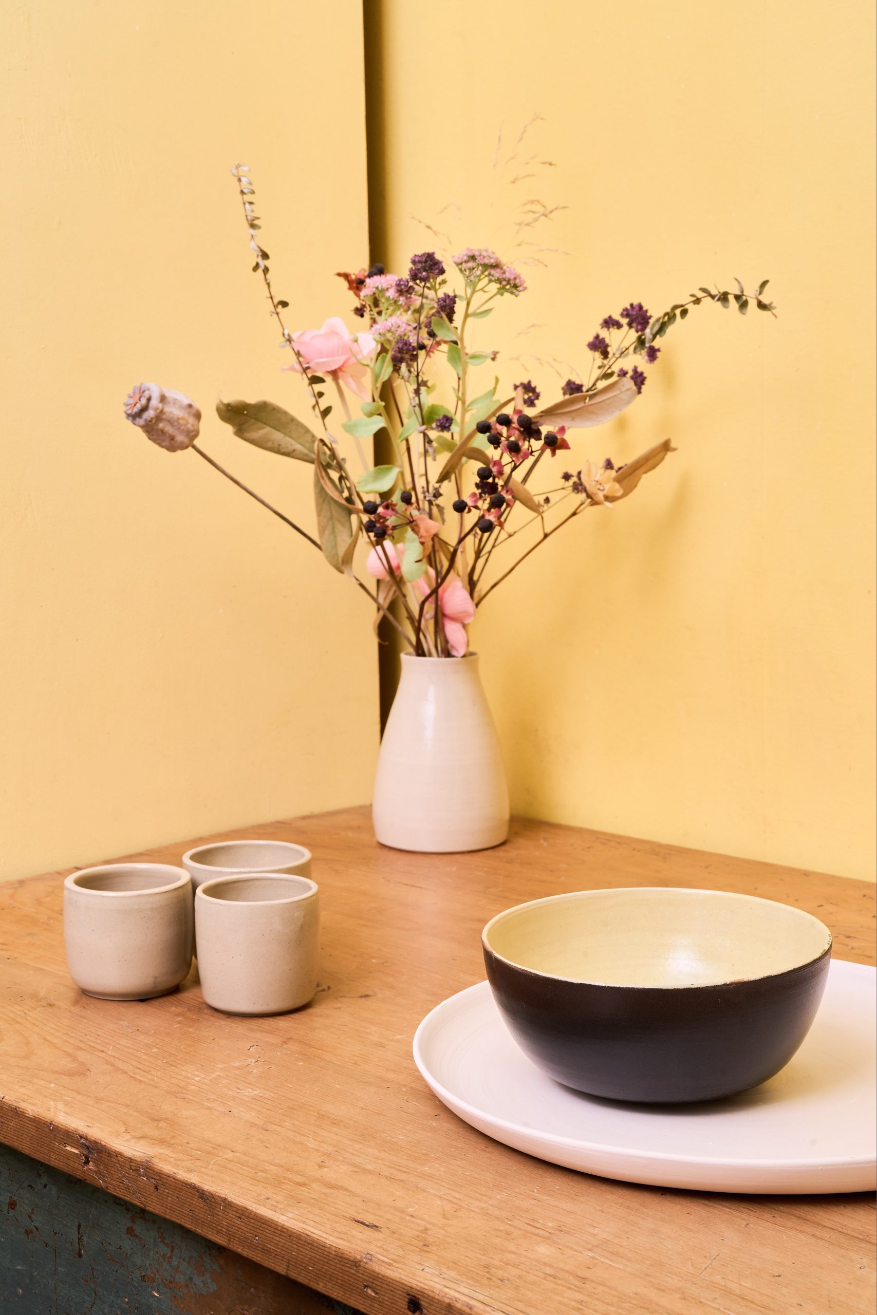 Handgefertigte Keramik: beige Becher, Vase mit Trockenblumen und zweifarbige Schale auf Holztisch – modern und minimalistisch arrangiert.